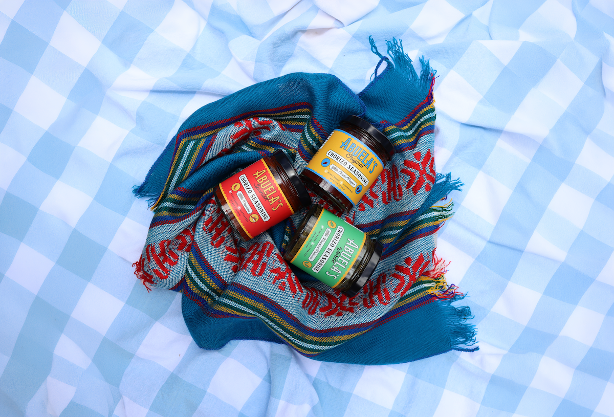 Three jars of gourmet spreads or cheeses arranged on a blue and red patterned woven blanket over a light blue and white checkered fabric background.