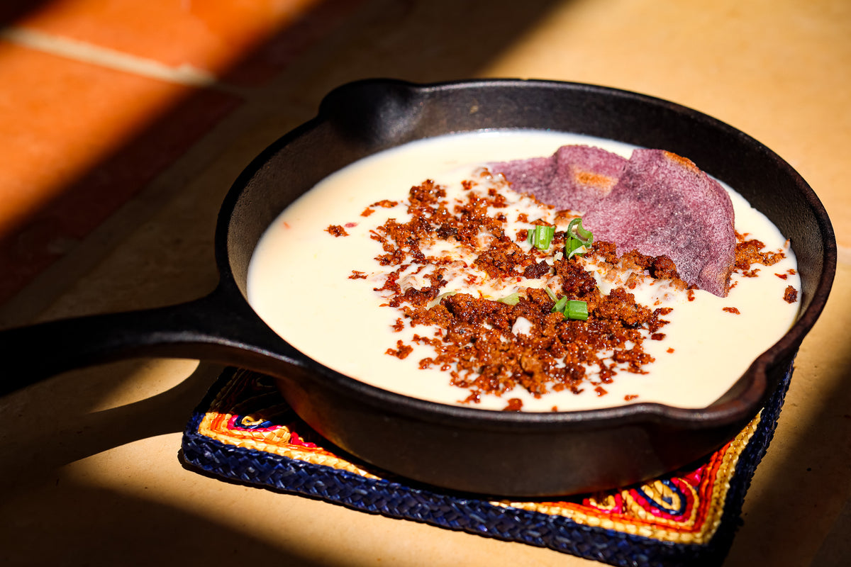 A black cast iron skillet filled with creamy white soup or mole, topped with coarse brown seasoning, a purple chip, and small green herb pieces, resting on a patterned blue and red cloth over a warm tan surface.