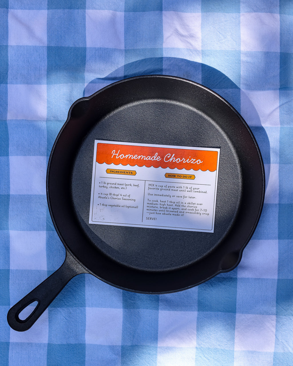 Overhead view of a cast iron skillet with the Homemade Chorizo recipe card inside, on a gingham cloth