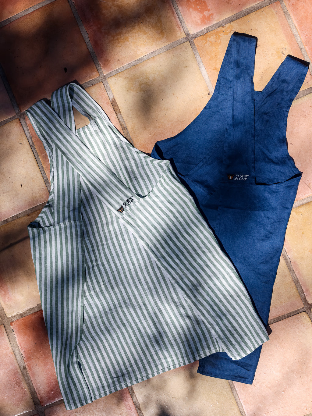 Blue and Green Stripe linen aprons laid flat side by side on terracotta tile