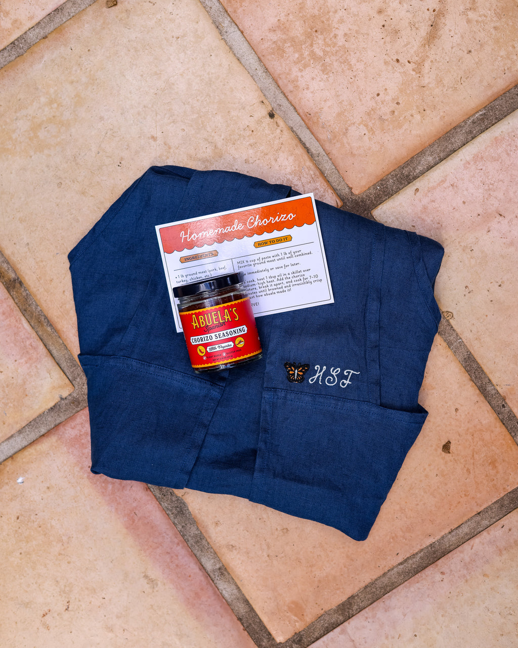 Folded Blue linen apron with the recipe card and a jar of Spanish chorizo seasoning on terracotta tile