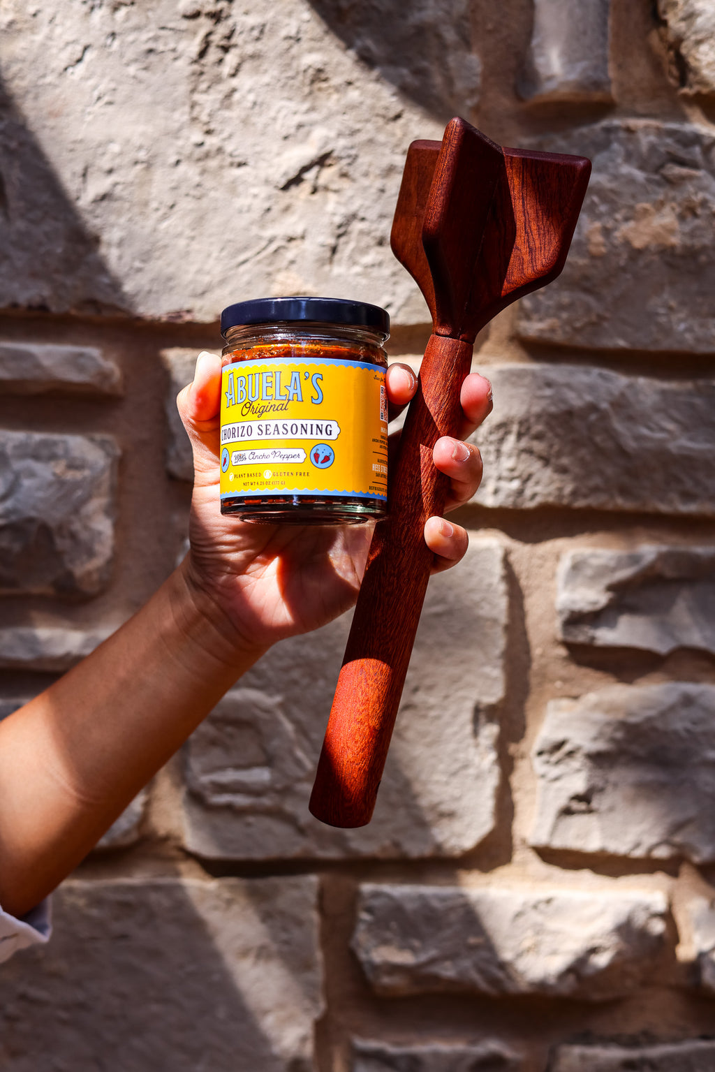 Raised hand holding a jar of Original chorizo seasoning and a teak chopper against a stone wall