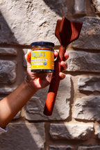 Raised hand holding a jar of Original chorizo seasoning and a teak chopper against a stone wall