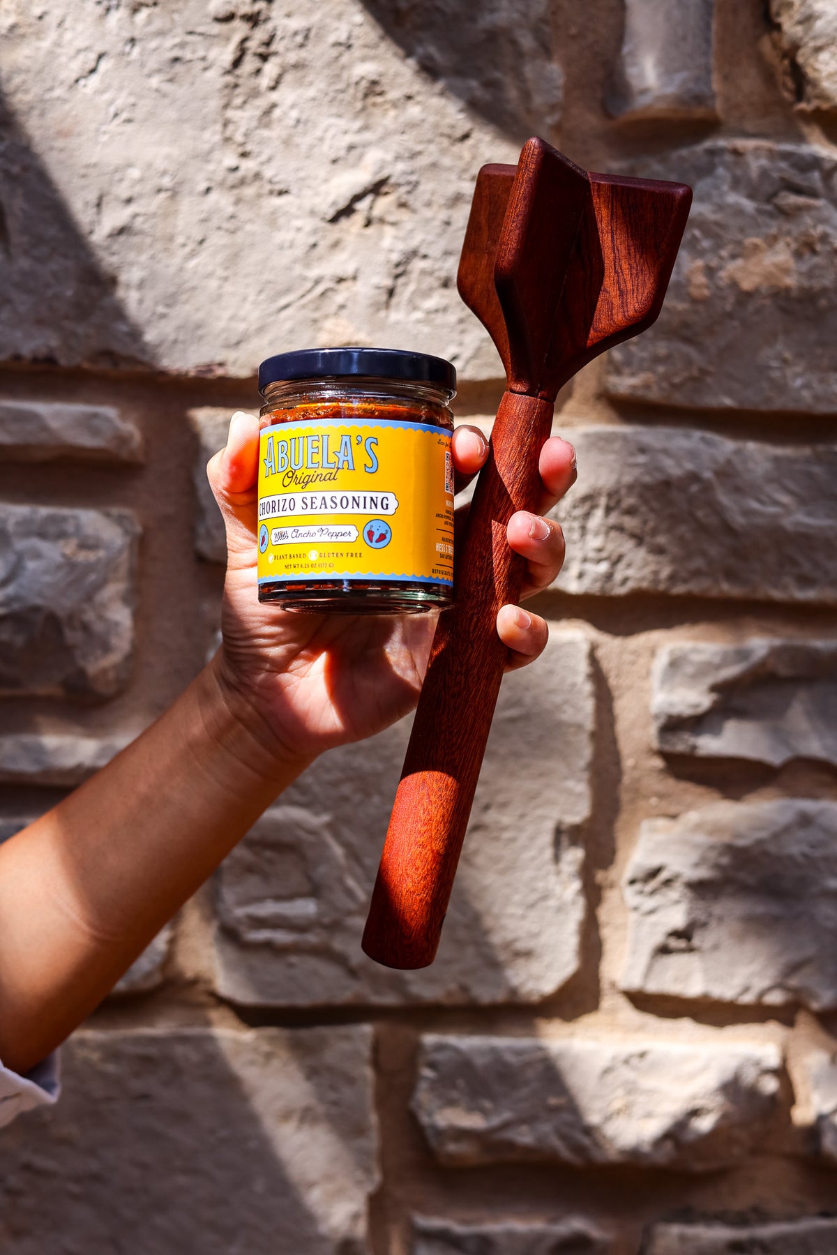 Raised hand holding a jar of Original chorizo seasoning and a teak chopper against a stone wall