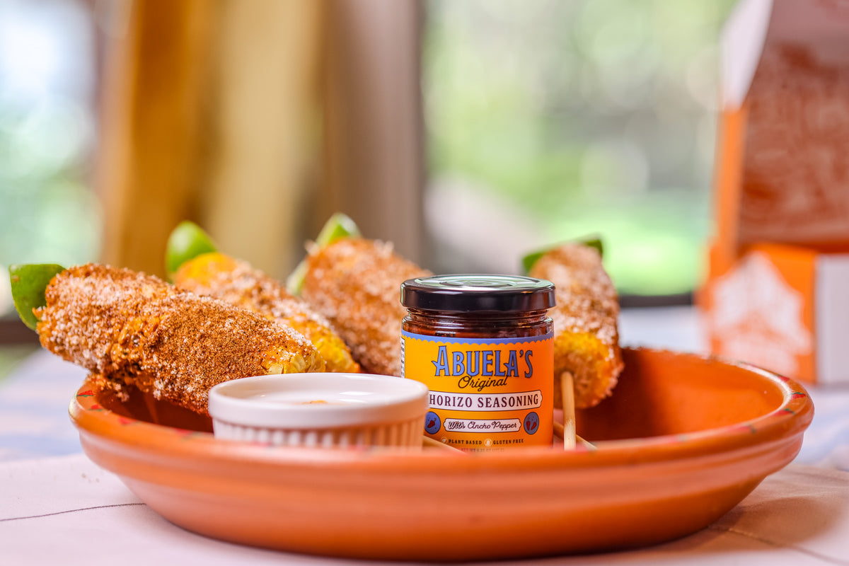 A small jar of Abuela's Guadizo Seasoning sits on a terracotta plate with corn on the cob and a small dipping cup, set in a softly blurred indoor environment.