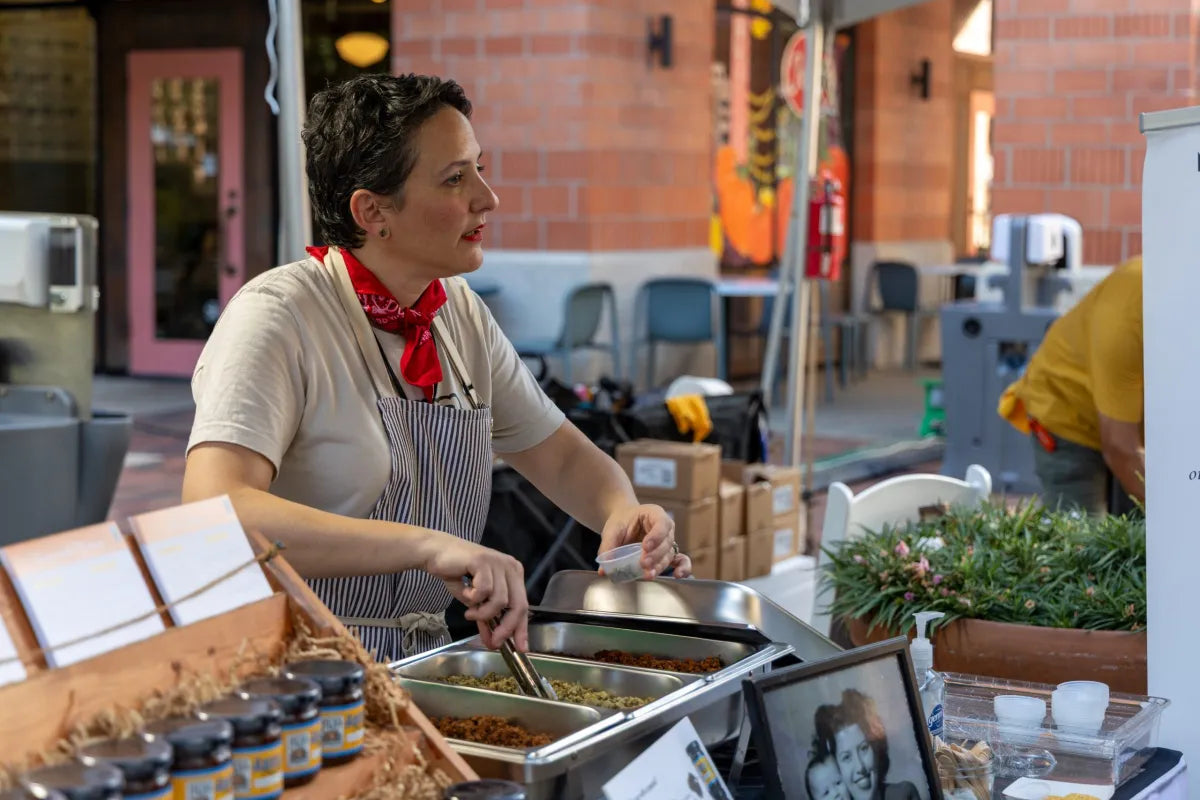 Woman working at an outdoor market stall with food and products.