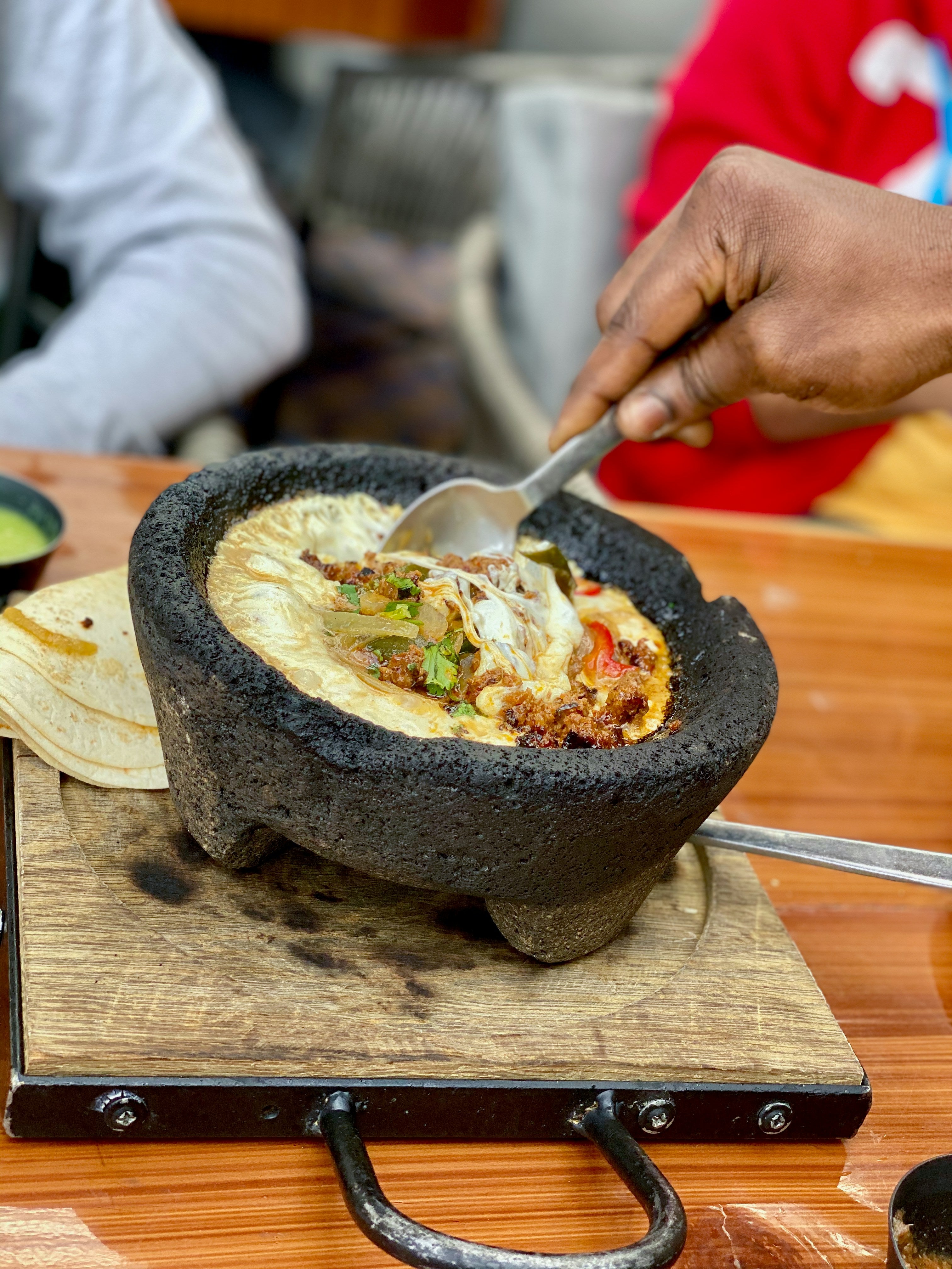 Person using a spoon to eat from a molcajete filled with food on a wooden surface.